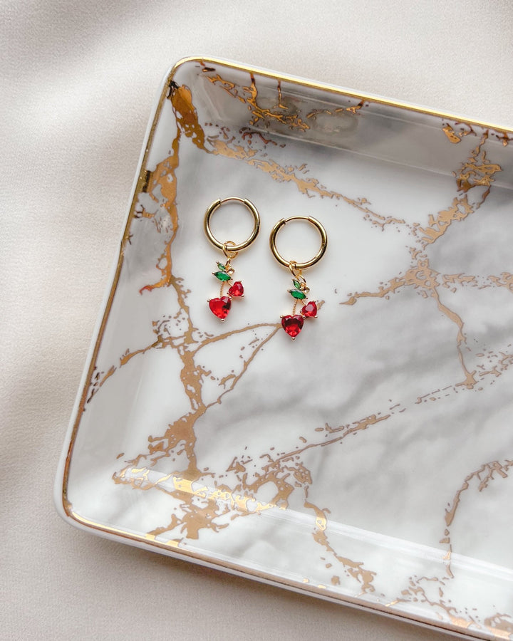 Boucles d'oreilles cerises posées sur une assiette blanche et dorée faisant ressortir le rouge intense des cerises.